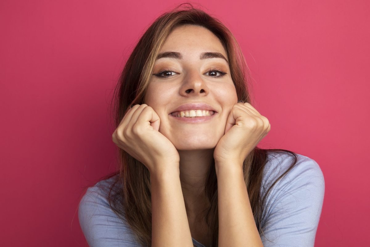 young beautiful woman in blue t-shirt looking at camera leaning head on fists happy and positive smiling standing over pink background