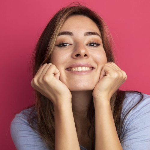 young beautiful woman in blue t-shirt looking at camera leaning head on fists happy and positive smiling standing over pink background